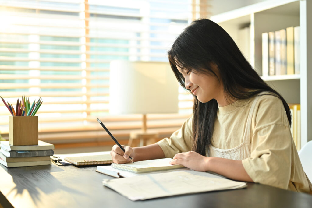 Middle-school student writing narration in a notebook at a sunlit desk with books and pencils.