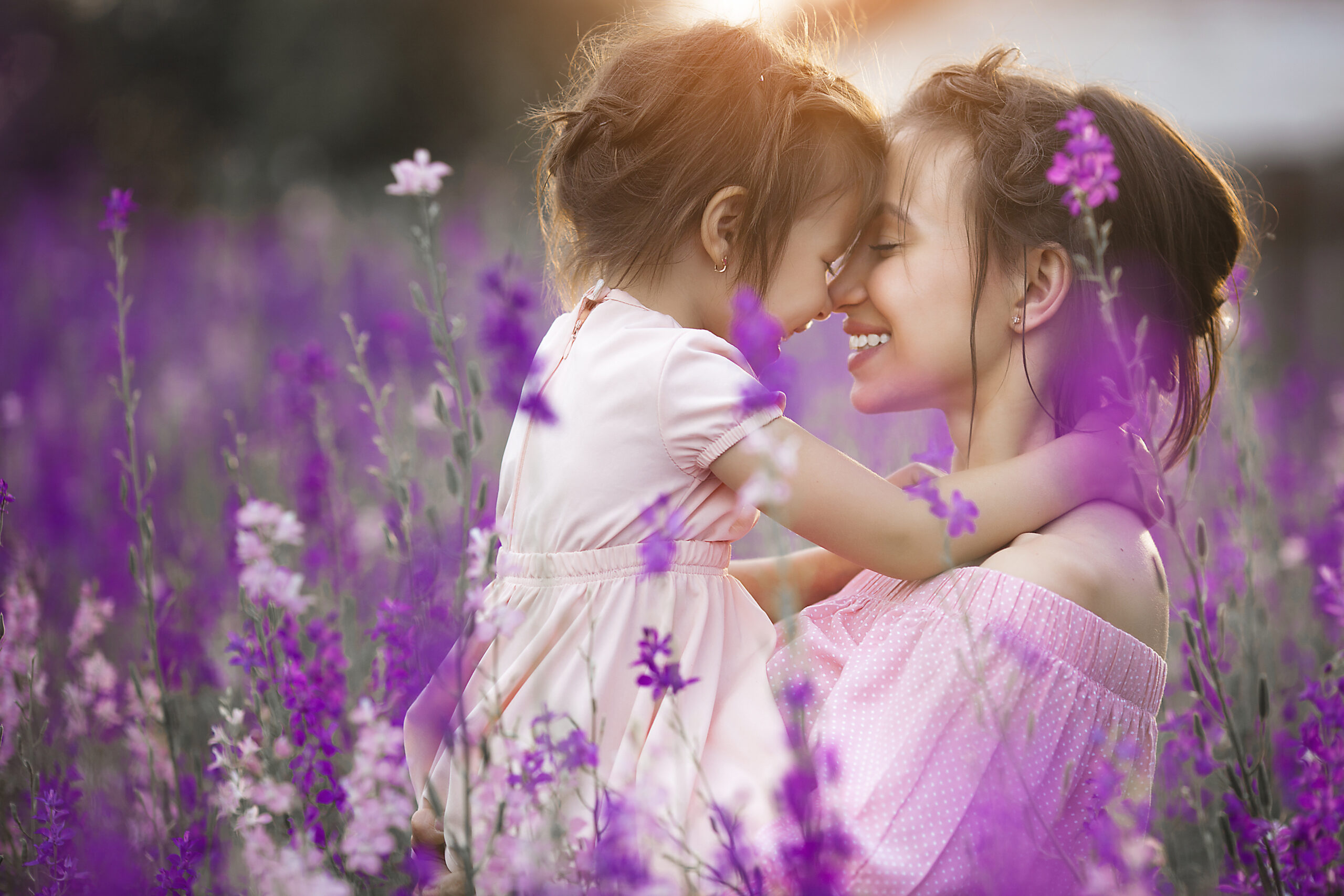 Mother and young daughter smiling forehead to forehead in a sunlit lavender field, a quiet moment of comfort and rest.