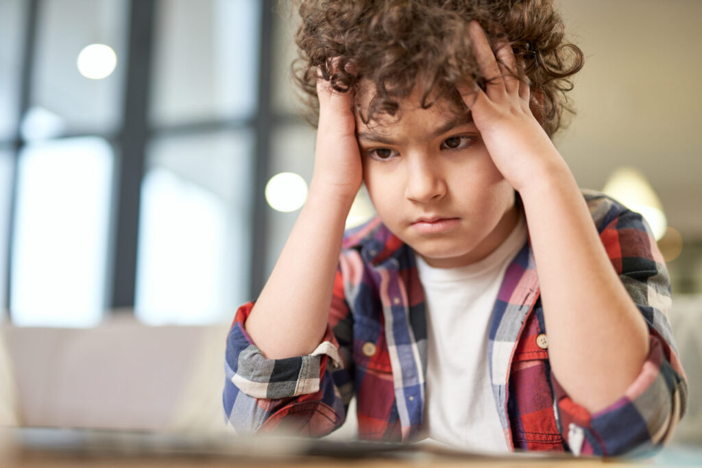 Child holding their head with both hands over schoolwork, looking discouraged in a softly lit home setting.