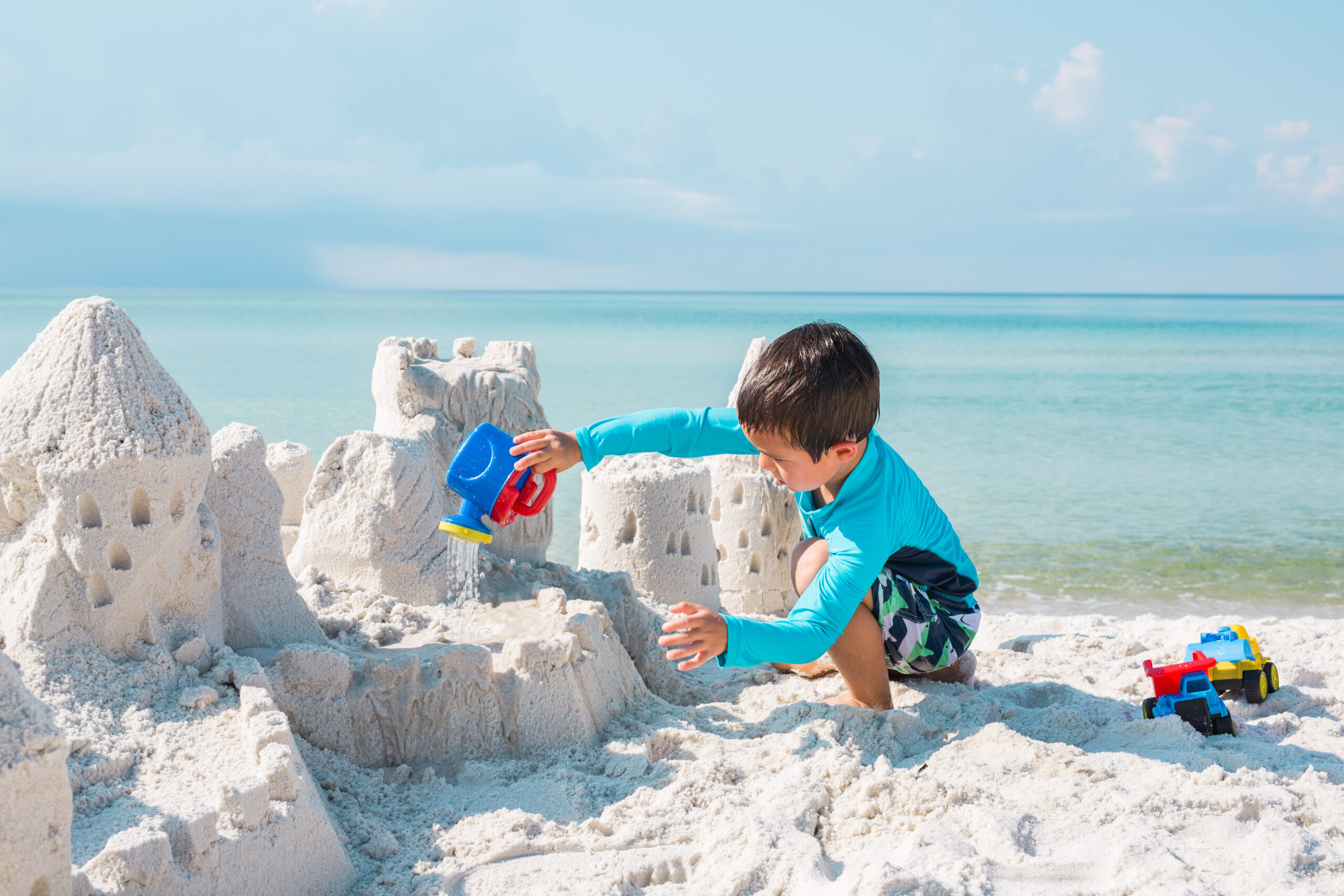 A young boy in a blue rashguard is intently using a red and blue watering can to finish a large, detailed sandcastle, illustrating the focused, joyful acquisition of grammar stage learning.
