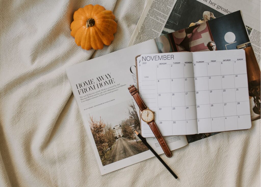 Open November monthly calendar with leather watch, pencil, magazine, and small pumpkin on a cozy blanket.