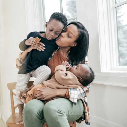 Young homeschool mother holding a toddler and baby by a bright window